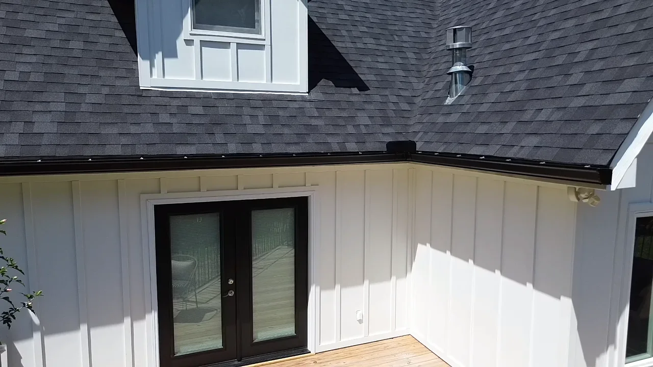 Dark seamless gutters with downspout installed on white fascia board beneath dark asphalt shingle roof